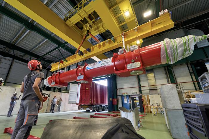cylindrical red structure lifted on yellow crane, with a man controlling it with a boxy remote control